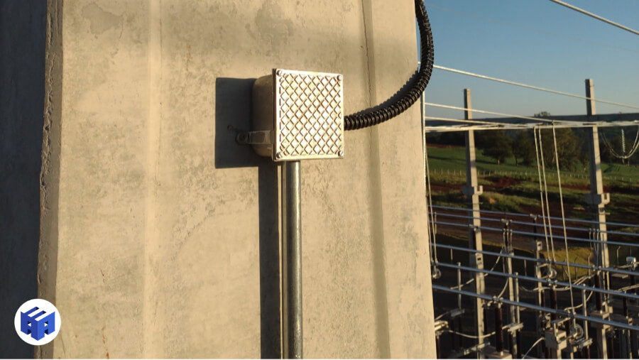 Imagem de um eletroduto fixado na parede externa de uma estrutura, com um conduíte corrugado conectando-o a outros componentes em um ambiente industrial. A tubulação, que protege os cabos internos contra danos físicos e interferência eletromagnética, está posicionada ao lado de uma subestação elétrica, evidenciando seu uso em locais que exigem proteção robusta para instalações elétricas.