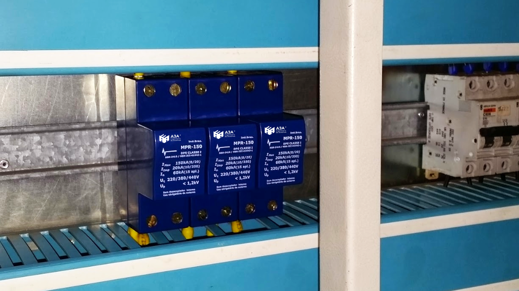 A imagem mostra um painel elétrico com três dispositivos de proteção contra surtos (DPS) azuis montados em um trilho.